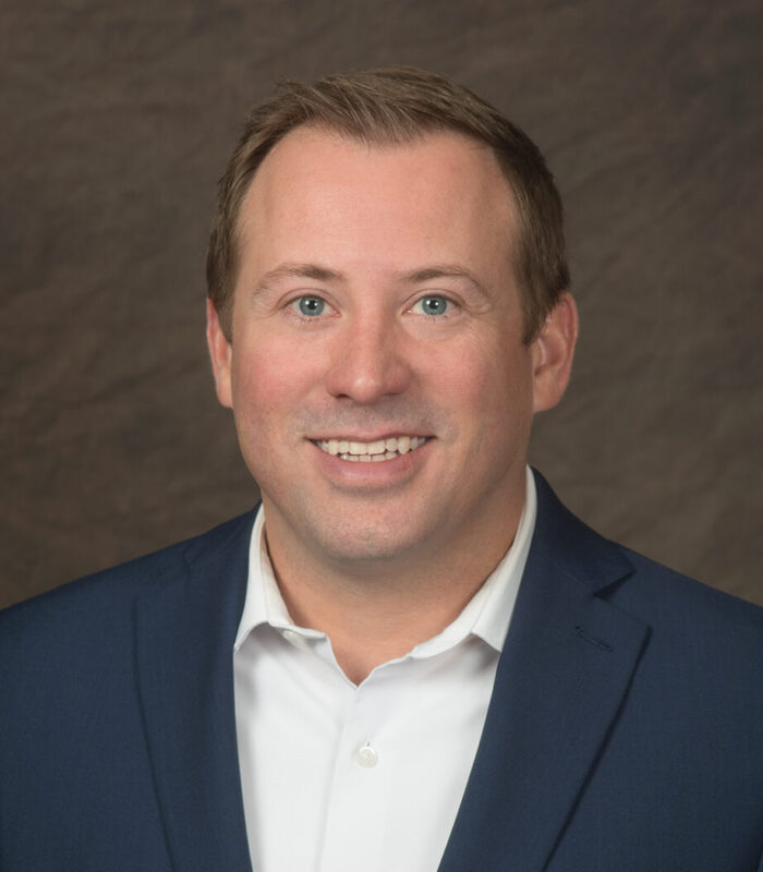 Headshot portrait of Mike Bellmore with short light brown hair and blue eyes, wearing a dark navy blazer over an open-collar white dress shirt, smiling against a textured brown backdrop.